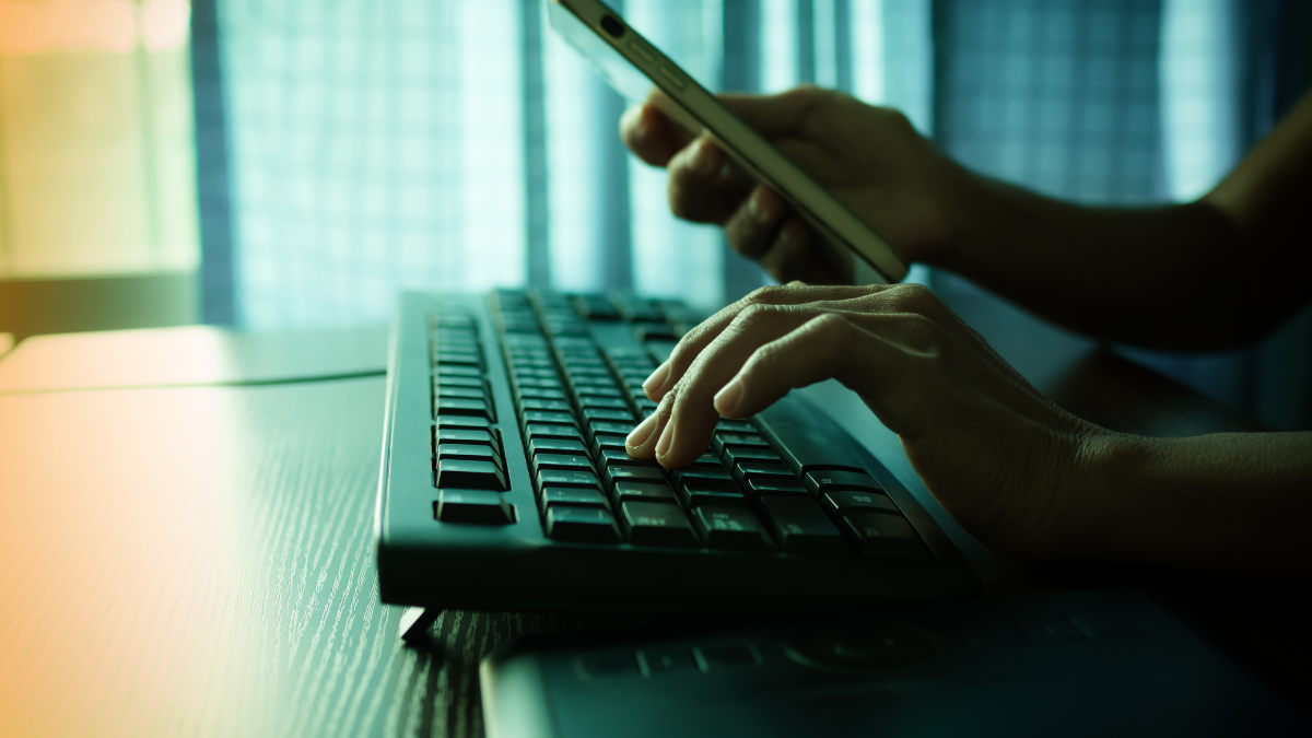 Close-up of hands typing on a keyboard while holding a smartphone, symbolizing online transactions, digital activity, and cyber operations.