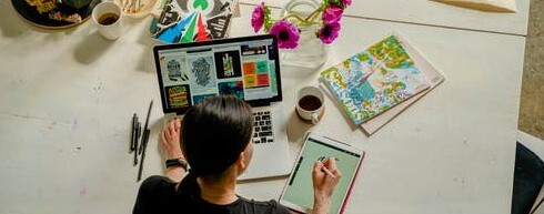Woman draws on a graphics tab, sat at her desk with coffee laptop sketchbooks and drawing equipment.