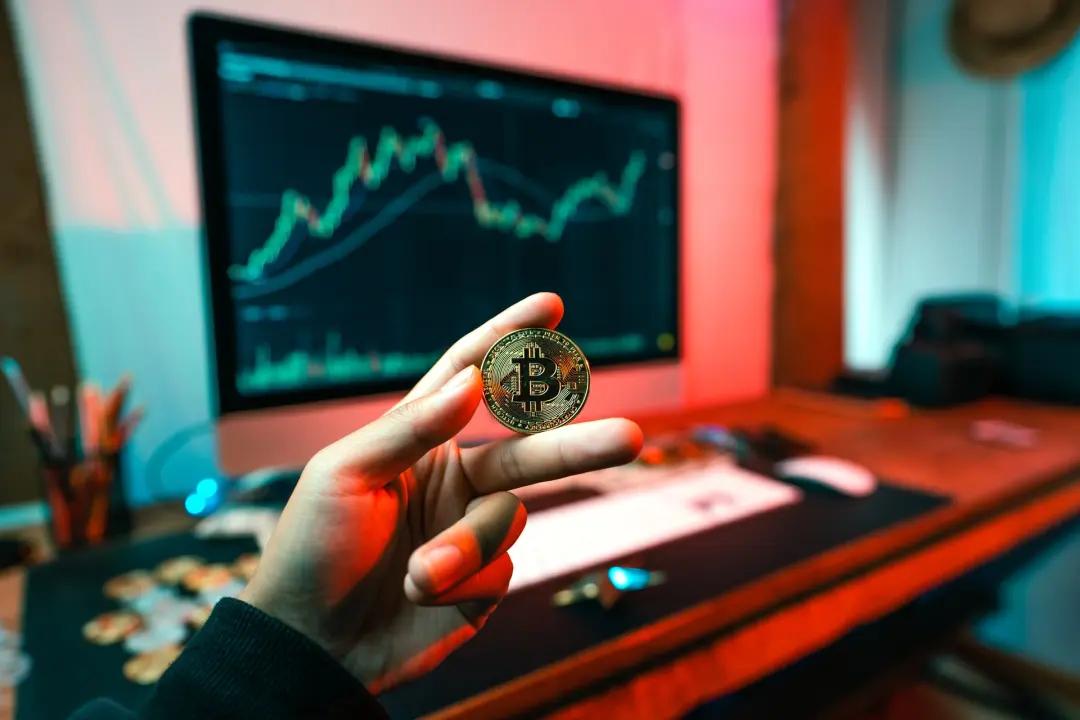Man holding up physical Bitcoin in front of a computer monitor